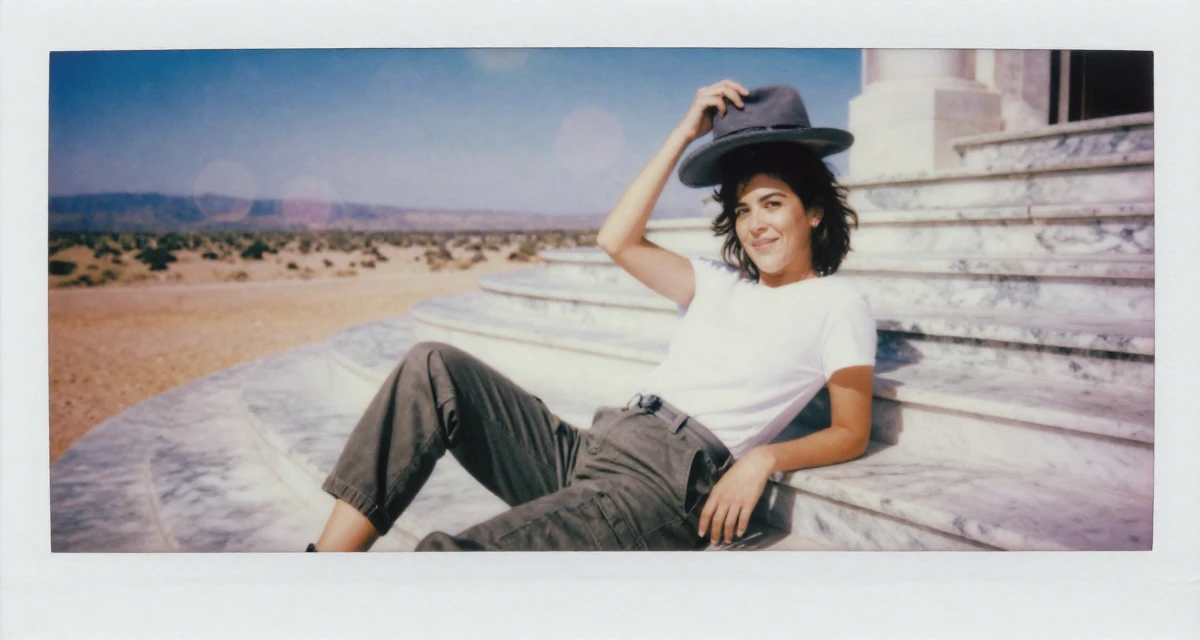 A amused Female From Australia, has a diploma in business administration in their 24, learning to prioritize emotional well-being,
wearing a simple t-shirt and cargo pants, holding a hat in a desert landscape.
