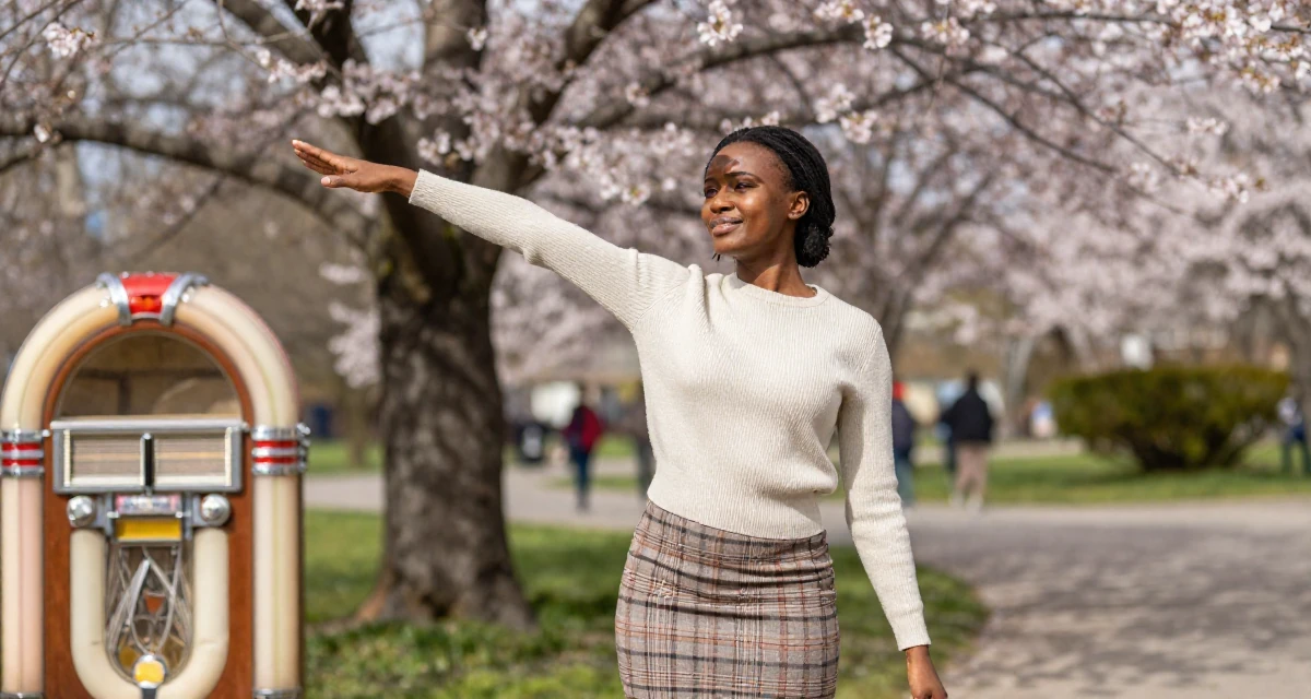 A radiant Female From Zambia, majored in marketing communication in their 23, discovering the allure of slow, controlled movement, wearing a fitted merino wool sweater and plaid mini skirt, shading eyes from the sun in a cherry blossom park.