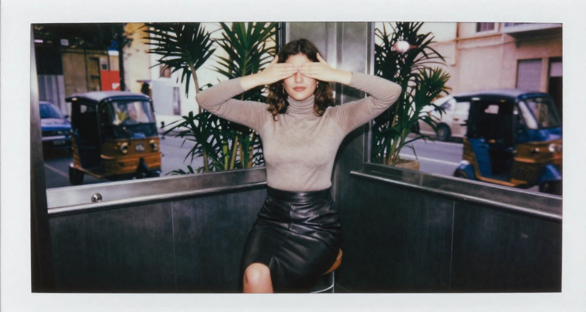 A calm Female From Athens Greece, learned movement expression through traditional dance in their 22, crafting content at midnight after work shifts, wearing a fitted turtleneck and a leather midi skirt, covering eyes from the sun in a elevator hall.