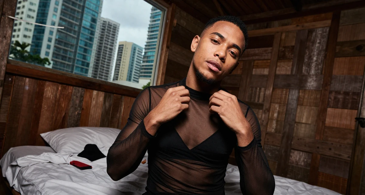 A quietly confident male From Trinidad & Tobago, studied performing arts in their 25, upgrading equipment slowly as earnings grow, wearing a sheer mesh top layered over a black bralette, fixing a collar in a rustic wooden cabin interior.