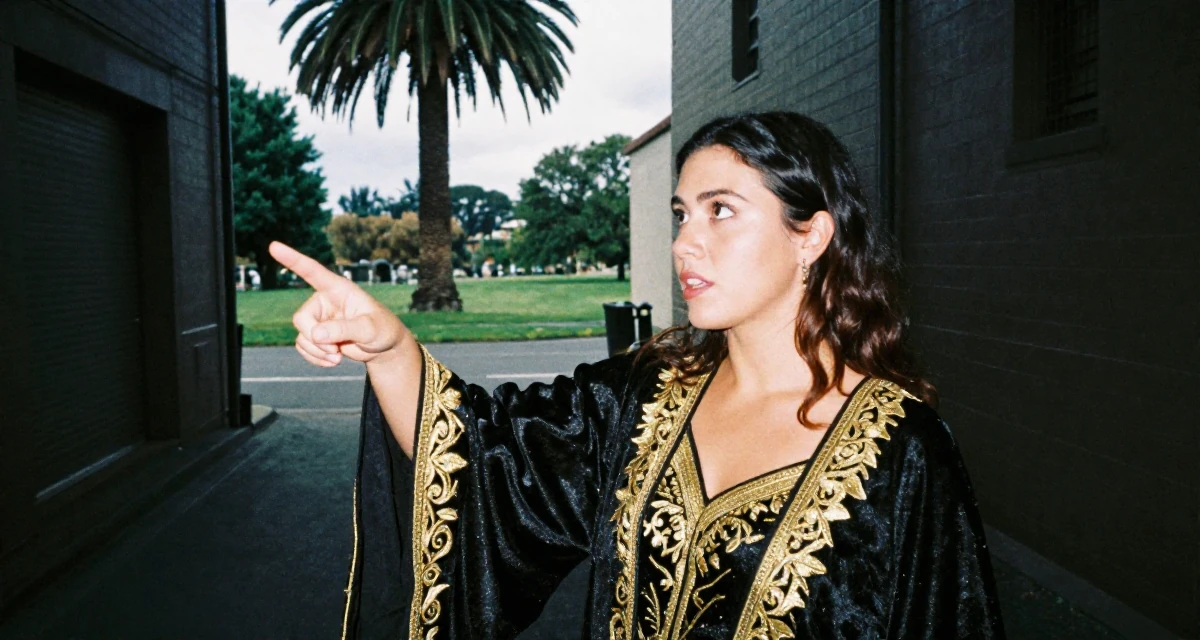 A intense and captivating Female From Wellington New Zealand, majored in sociology in their 25, discovering early creative passions, wearing a high-fantasy sorceress robe with gold embroidery, pointing at something nearby in a quiet alleyway.