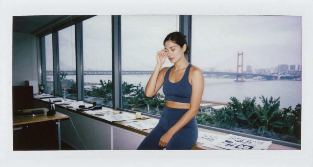 A calm Female Once a recruitment assistant, now creating relatable work-life content in their 36, expert in interior design and home decor, wearing a form-fitting athletic set with a sports bra and leggings, rubbing eyes in a glass-walled conference room.